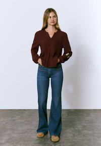 Blouse texturée bordeaux avec un col en V et des manches longues, associée à un jean bleu évasé et des chaussures beiges sur un sol gris.