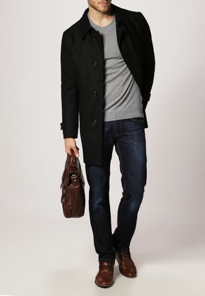 Cappotto lungo nero, maglione grigio, jeans blu e scarpe di pelle marrone. Tiene una borsa di pelle marrone. Design semplice, vestibilità su misura.