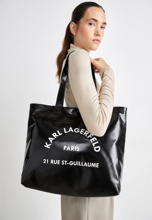Kvinde med langt hår holder en stor sort shopper med hvid tekst "Karl Lagerfeld Paris 21 Rue St-Guillaume" på skulderen.