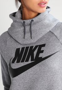 Felpa grigia con cappuccio, caratterizzata da un grande logo nero "NIKE" e lacci con terminazioni in argento. Tessuto liscio, design sportivo.