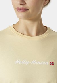 Sv�ětle žluté bavlněné tričko s nápisem "Helly-Hansen" v bílém písmu a malou norskou vlajkou. Design s kulatým výstřihem a hladkou texturou.