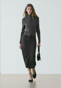 Grauer, gerippter Reißverschluss-Pullover, schwarzer Midirock, beige Socken und schwarze Ballerinas. Modell hält eine kleine schwarze Tasche und steht auf einer glatten Oberfläche.