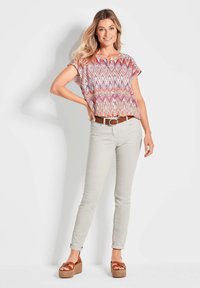 Gemustertes weißes T-Shirt in Pink und Blau, kombiniert mit hellgrauen, enganliegenden Jeans und braunen Keil-Sandalen. Ein brauner Gürtel rundet das Outfit ab.