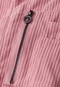 Primo piano di un tessuto a coste rosa con una zip verticale nera dotata di un cursore in metallo rotondo e di una piccola tasca accanto.