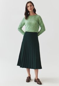 Maglione verde texture abbinato a una gonna plissettata verde scuro, completato da scarpe slip-on marroni; posizionato su uno sfondo bianco semplice.