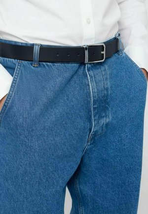 Jeans in denim blu con vestibilità comoda, caratterizzati da cuciture a contrasto e tasche laterali, abbinati a una cintura in pelle nera e una camicia bianca.
