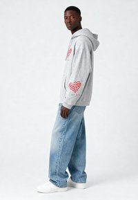 Sudadera gris con patrones de corazones rojos en la parte frontal y las mangas, combinada con jeans de corte suelto de color azul claro y zapatillas blancas.
