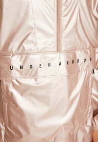 Jasnoróżowa kurtka Under Armour z zamkiem błyskawicznym, pionową prążkowaną fakturą i czarnymi napisami na plecach. Gładki, błyszczący materiał.