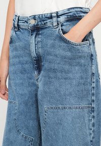 Gros plan de jeans en denim bleu avec grandes poches avant, une main dans la poche, fermeture visible par bouton métallique et passants de ceinture.