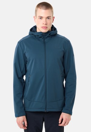 Giovane uomo visto di fronte che indossa una giacca con cappuccio blu con cerniere e tasche con zip, e pantaloni neri, su uno sfondo semplice.