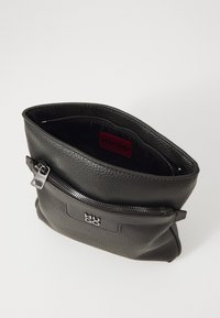 Črna usnjena torbica z teksturirano površino. Ima zadrgo na vrhu in sprednjo žepno z detajlom logotipa. Notranjost je obložena s črno tkanino.