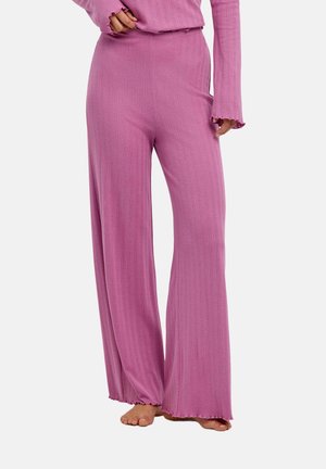 Femme portant un pantalon rose large côtelé et un haut à manches longues assorti, debout pieds nus sur un fond uni.