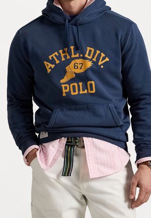 Homme portant un sweat à capuche bleu marine avec texte jaune "ATHL. DIV. 67 POLO" et motif chaussure ailée, chemise rose rayée, pantalon blanc et ceinture rayée.