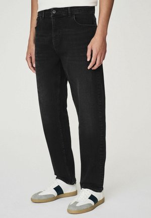 Homme portant un jean noir droit et des baskets blanches avec des accents gris et bleu foncé, debout devant un fond uni.