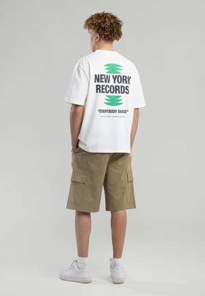 White short-sleeve t-shirt with black and green graphic text on back, paired with olive green cargo shorts and white sneakers.