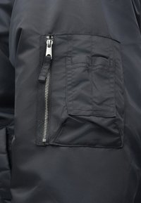 Μαύρο ύφασμα για μπουφάν με κοντινή λήψη από μια φερμουάρ τσέπη και μια επίπεδη τσέπη. Διαθέτει λεία υφή και μινιμαλιστικά σχεδιαστικά στοιχεία.