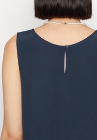 Takakuvasta kuvatut henkilön, jolla on lyhyet tummat hiukset, päällä on ilman hihoja oleva tummansininen toppi, jossa on yksi takanappi, sekä helmikaulakoru.