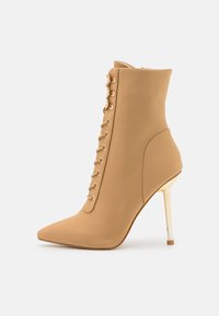 Bottine beige à bout pointu, en tissu extensible, avec fermeture éclair. Éléments métalliques à lacets et talon stiletto doré brillant.