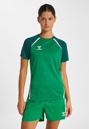 Femme debout, face à l'avant, portant un maillot de sport Hummel vert avec des manches vert foncé et un short assorti, sur un fond blanc.
