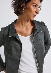 Vrouw met kort krullend haar, die zilveren hoopoorbellen, een witte shirt en een open donkergrijze jas draagt, en naar de zijkant kijkt tegen een effen achtergrond.