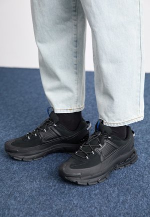 Des chaussures de sport noires avec un tissu texturé et des semelles en caoutchouc, dotées de lacets élastiques et d'un accent bleu au talon. Portées avec un pantalon en denim clair.