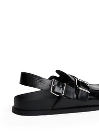 Sandalo in pelle nera con cinturini regolabili a fibbia, suola piatta e superficie liscia. Presenta un design minimalista e una forma arrotondata.