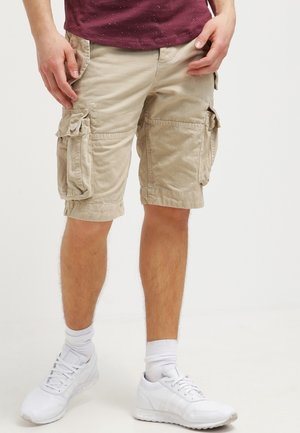 Mand iført beige cargoshorts, hvide sokker og hvide sneakers, stående mod en ensfarvet baggrund.