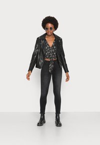 Jaqueta de motociclista de couro preto sobre um top cropped estampado, combinada com jeans skinny pretos e botas de tornozelo pretas. Óculos de sol redondos completam o visual.