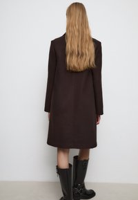 Cappotto in lana marrone scuro con maniche lunghe, taglio dritto e dettagli minimi. Indossato con stivali in gomma neri alti fino al ginocchio.