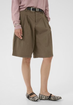 Kvinde iført brune knælange plisserede shorts, lyserød bluse med lange ærmer, sort bælte, sølvarmbånd og zebra-stribede flade sko.