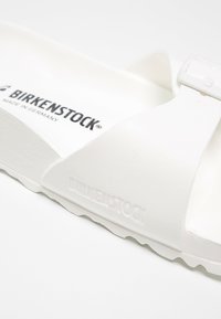 Gros plan sur une bride et une semelle intérieure blanches d'une sandale Birkenstock montrant le logo en relief et le texte de la marque imprimé en noir.