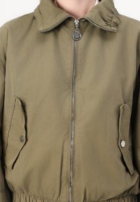 Giacca leggera verde oliva con colletto alto, chiusura frontale con zip e due tasche a pattina con bottoni a scatto. Vita leggermente arricciata.