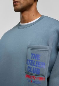 Lichtblauwe sweatshirt met ronde hals, voorzien van een voorkantzak en tekst in blauw en rood, gemaakt van zachte, Textuurstof.