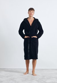 Accappatoio nero con cappuccio realizzato in materiale morbido e testurizzato, caratterizzato da una vita con cintura, ampie tasche frontali e nastro con logo in rilievo lungo la parte anteriore.