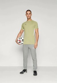 Muž stojící s fotbalovým míčem značky Puma, oblečený v světle zeleném tričku Puma, šedých teplácích a černých sportovních botách.