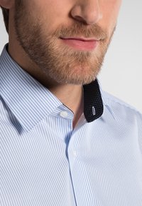 Camicia a righe azzurro chiaro con colletto nero decorato da pois bianchi, chiusura con bottoni e taglio sartoriale che mette in risalto il décolleté.