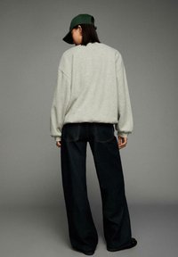 Graues, übergroßes Sweatshirt mit Fallärmeln, kombiniert mit weiten, dunklen Jeanshosen. Eine grüne Mütze rundet den Look ab.