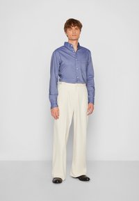 Blaues Hemd mit Knopfleiste und kleiner Brusttasche, kombiniert mit beigen, gefalteten Hosen; schwarze Schuhe runden das Outfit ab.