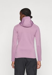 Sudadera de manga larga con capucha en color púrpura claro, con un diseño ajustado, textura suave y costuras en contraste, combinada con pantalones oscuros.