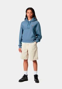 Sudadera con capucha de algodón azul claro con bolsillo tipo canguro, combinada con pantalones cortos crema y botas negras con cordones, sobre un fondo blanco.