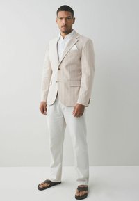 Helle beige gestreifte Jacke mit einem einzigen Knopf, kombiniert mit weißen Leinenhosen und schwarzen Flip-Flops, vor einem hellen Hintergrund.