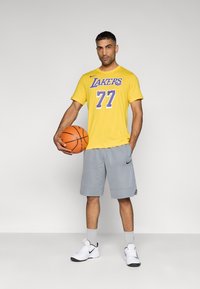 Κίτρινη φανέλα μπάσκετ με το "Lakers" και τον αριθμό "77" σε μωβ, συνδυασμένη με γκρι σορτς. Κρατάει μια μπάλα μπάσκετ, φοράει λευκά αθλητικά παπούτσια με μαύρες λεπτομέρειες.