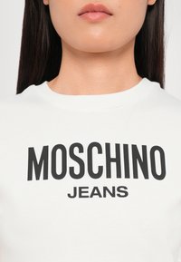 White cotton t-shirt featuring bold black lettering "MOSCHINO JEANS" across the chest. Smooth texture, crew neck design.
