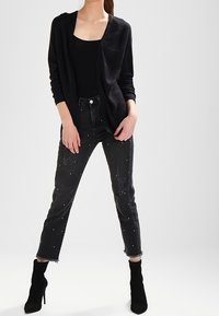 Czarny dziergany kardigan na czarnej bluzce, zestawiony z poszarpanymi czarnymi dżinsami ozdobionymi perłami i postrzępionymi nogawkami. Czarne botki za kostkę.