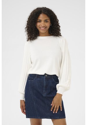 Femme aux cheveux bouclés souriante, portant un chemisier blanc plissé à manches longues et une jupe en denim bleu foncé, debout devant un fond uni.