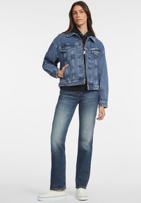 Denimska jakna v modri barvi z dolgimi rokavi, žepi na prsih, gumbi za zapiranje in sproščenim krojem, nizka z lahkimi modrimi kavbojkami in belimi supergami.