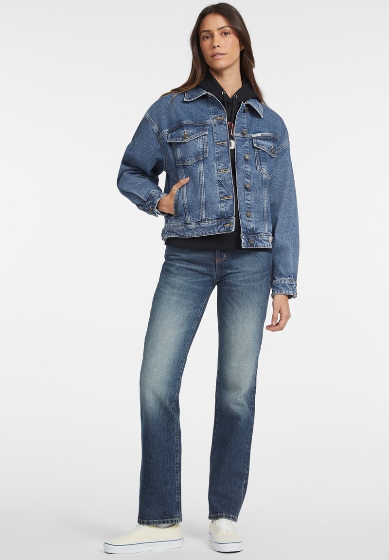Chaqueta de mezclilla azul con mangas largas, bolsillos en el pecho, cierres de botón y un corte relajado, combinada con jeans azul claro y zapatillas blancas.