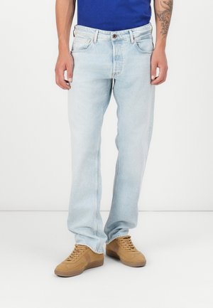 Mand iført lyseblå jeans, blå skjorte og beige sneakers står med hænderne afslappet ned langs siden mod en hvid baggrund.