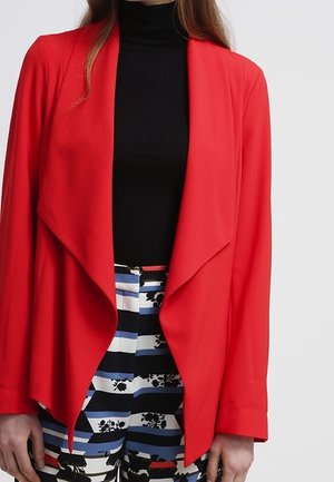 Nainen pukeutuneena kirkkaan punaiseen avoimeen bleiseriin mustan poolokauluksen ja sinivalkomustapunaruudulliset kukkakuviolliset housut päällään.