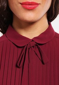 Blouse bordeaux avec un design plissé, un col et un détail à nouer à l'avant. Tissu léger avec une texture lisse.